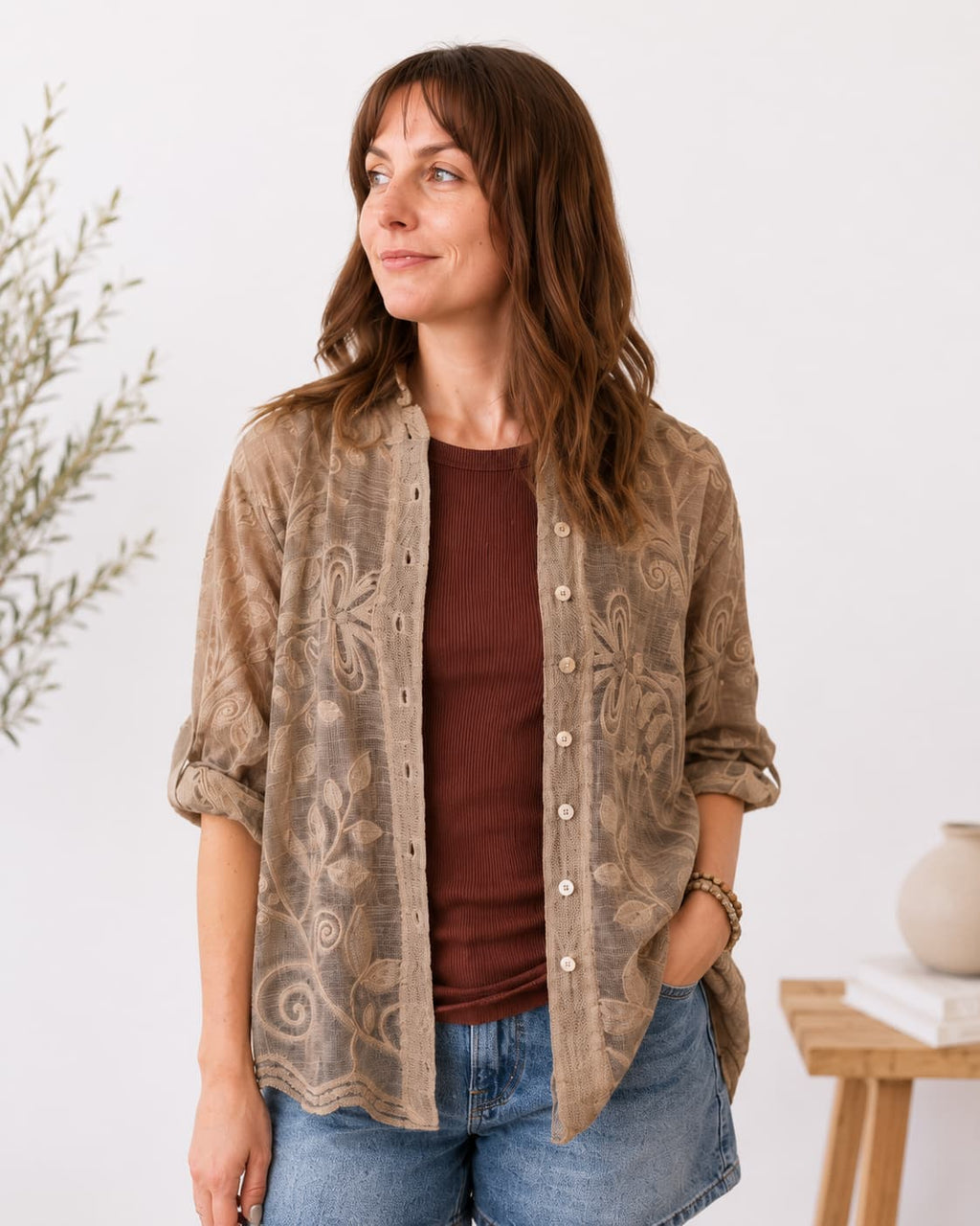 A woman wearing a brown bohemian-style sheer embroidered button-up shirt over a dark brown ribbed top and blue jeans, standing in a brightly lit indoor setting.