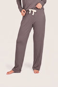 A person wearing loose, straight-leg lounge pants in a muted taupe-gray color with a contrasting cream drawstring, standing barefoot against a plain light beige studio background.