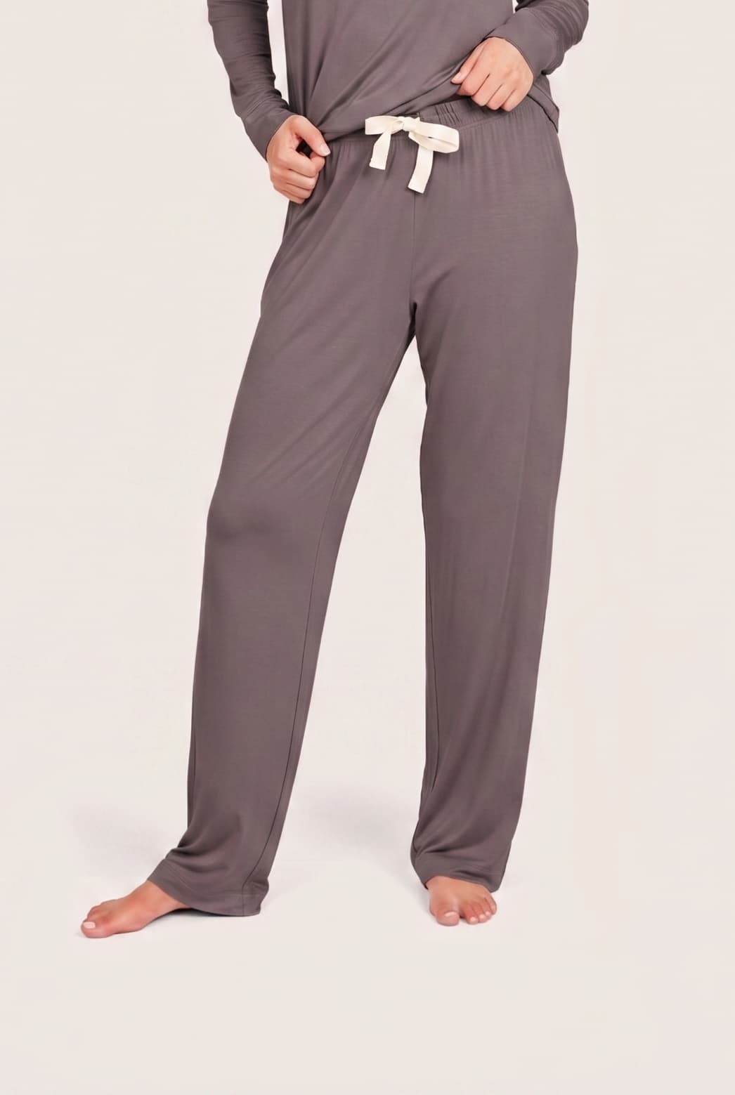 A person wearing loose, straight-leg lounge pants in a muted taupe-gray color with a contrasting cream drawstring, standing barefoot against a plain light beige studio background.