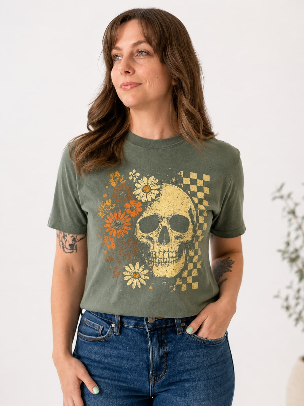 A woman wearing an olive green graphic t-shirt with a distressed yellow skull, orange and white flowers, and a checkered pattern, paired with blue jeans.