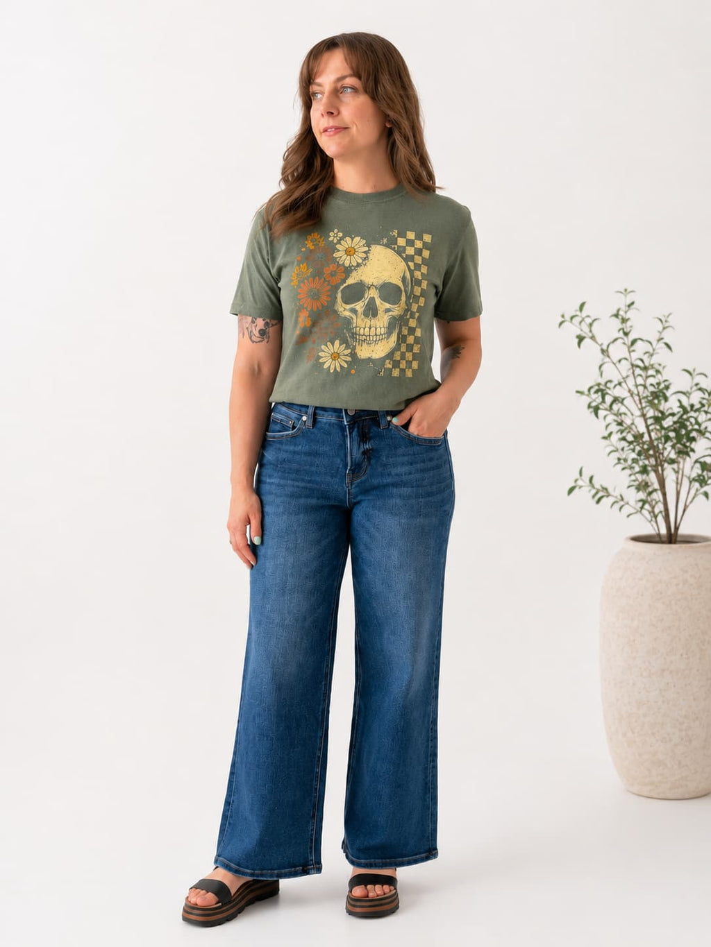 A woman modeling an olive green graphic t-shirt with a yellow skull and floral print, paired with wide-leg blue jeans and brown sandals, standing in a studio setting next to a potted plant.