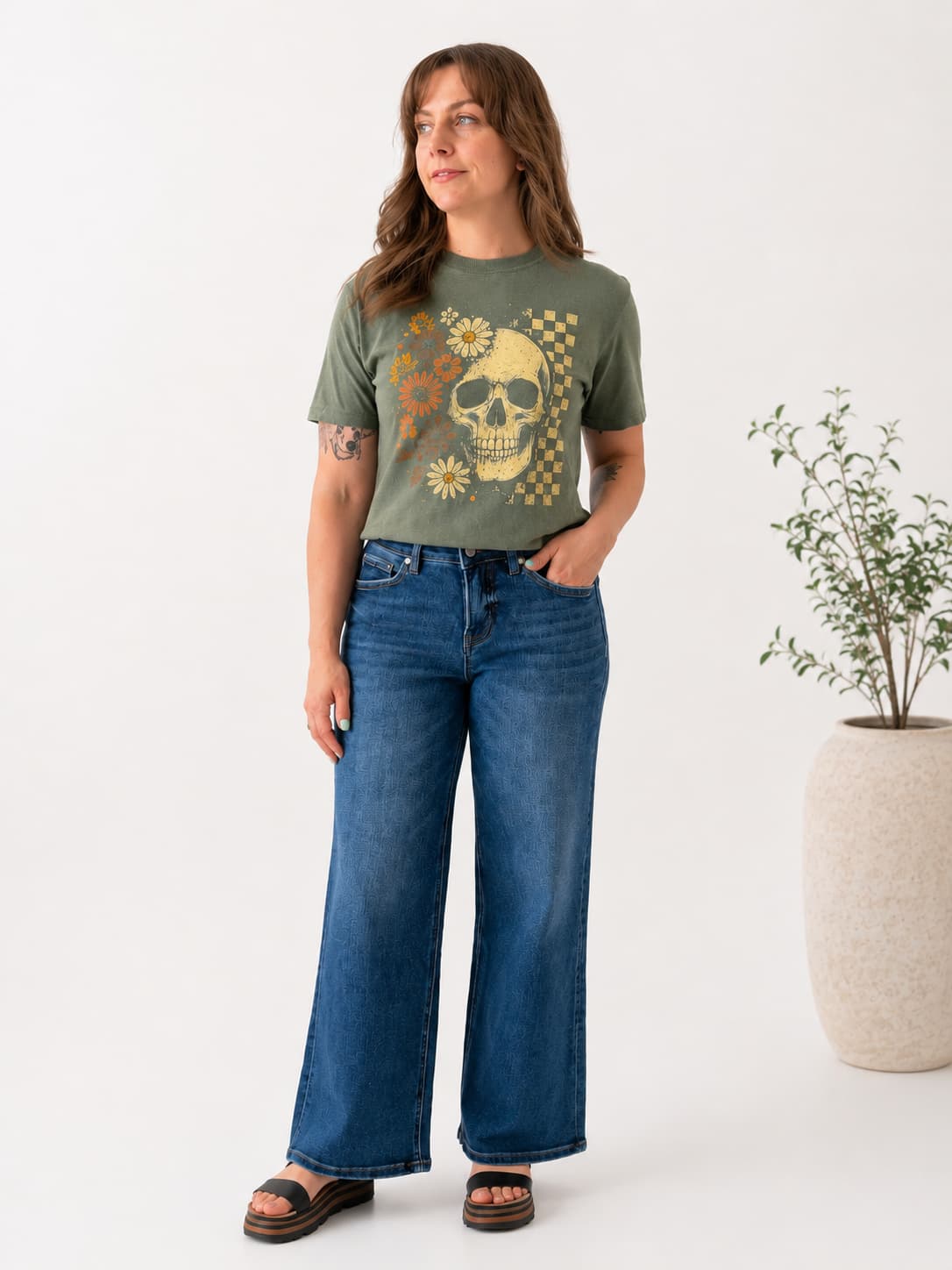 A woman modeling an olive green graphic t-shirt with a yellow skull and floral print, paired with wide-leg blue jeans and brown sandals, standing in a studio setting next to a potted plant.