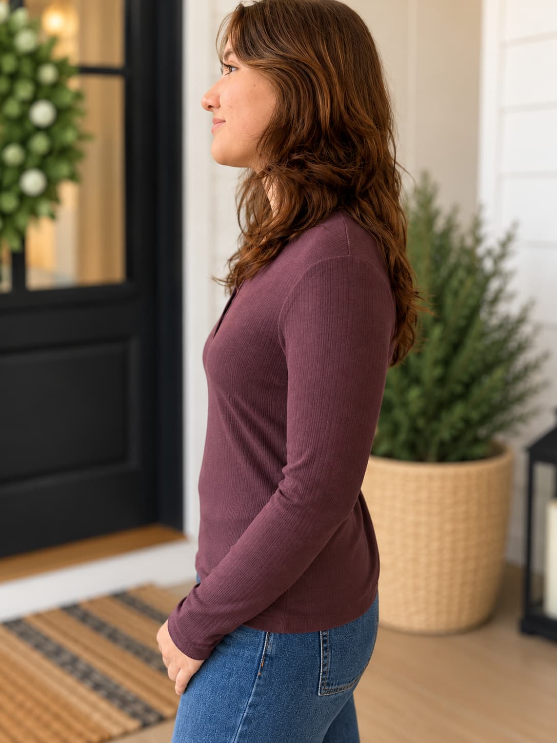 A woman in side profile wearing a ribbed long-sleeve henley top in a dusty plum purple color, paired with blue jeans, standing in a home entryway.