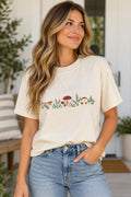 A woman wearing a cream-colored short-sleeve t-shirt with a colorful embroidered forest design across the chest, paired with light-wash blue jeans, standing in a bright, modern indoor setting.