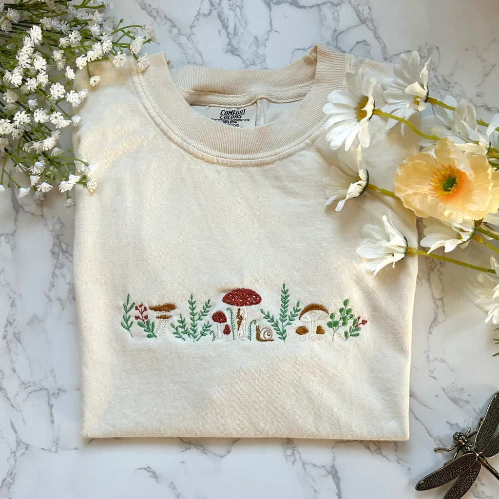 Beige sweatshirt with embroidered mushrooms and plants on a marble surface with flowers.