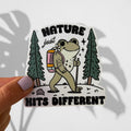 A hand with lavender nail polish holds a die-cut sticker featuring a cartoon frog hiking with a backpack and trekking pole, with the text 'NATURE just HITS DIFFERENT'. The sticker is shown against a white background with a leaf shadow.
