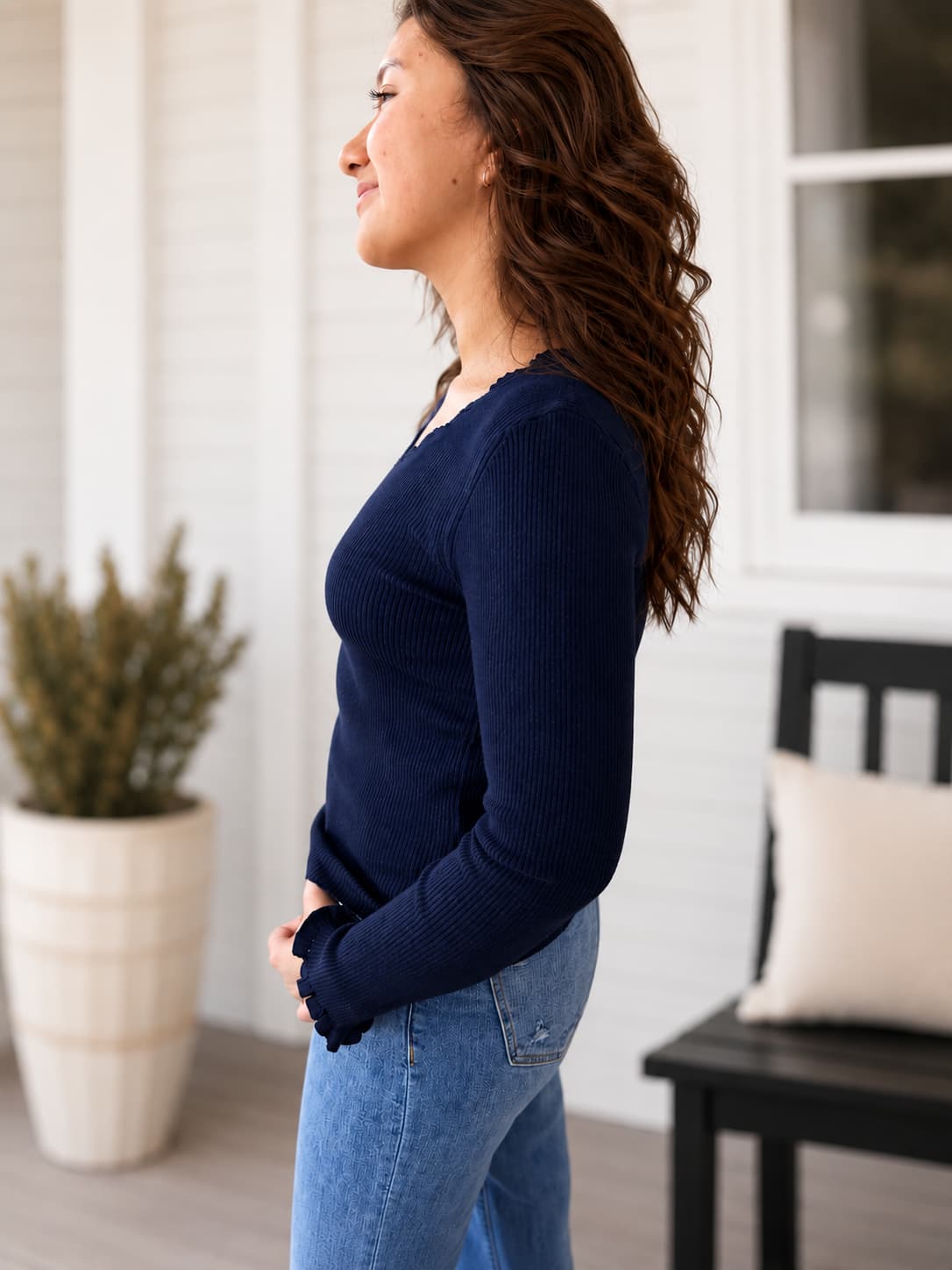 A woman wearing a navy blue ribbed long-sleeve sweater and light blue jeans, standing in profile on a porch with white siding, a potted plant, and a black bench in the background.