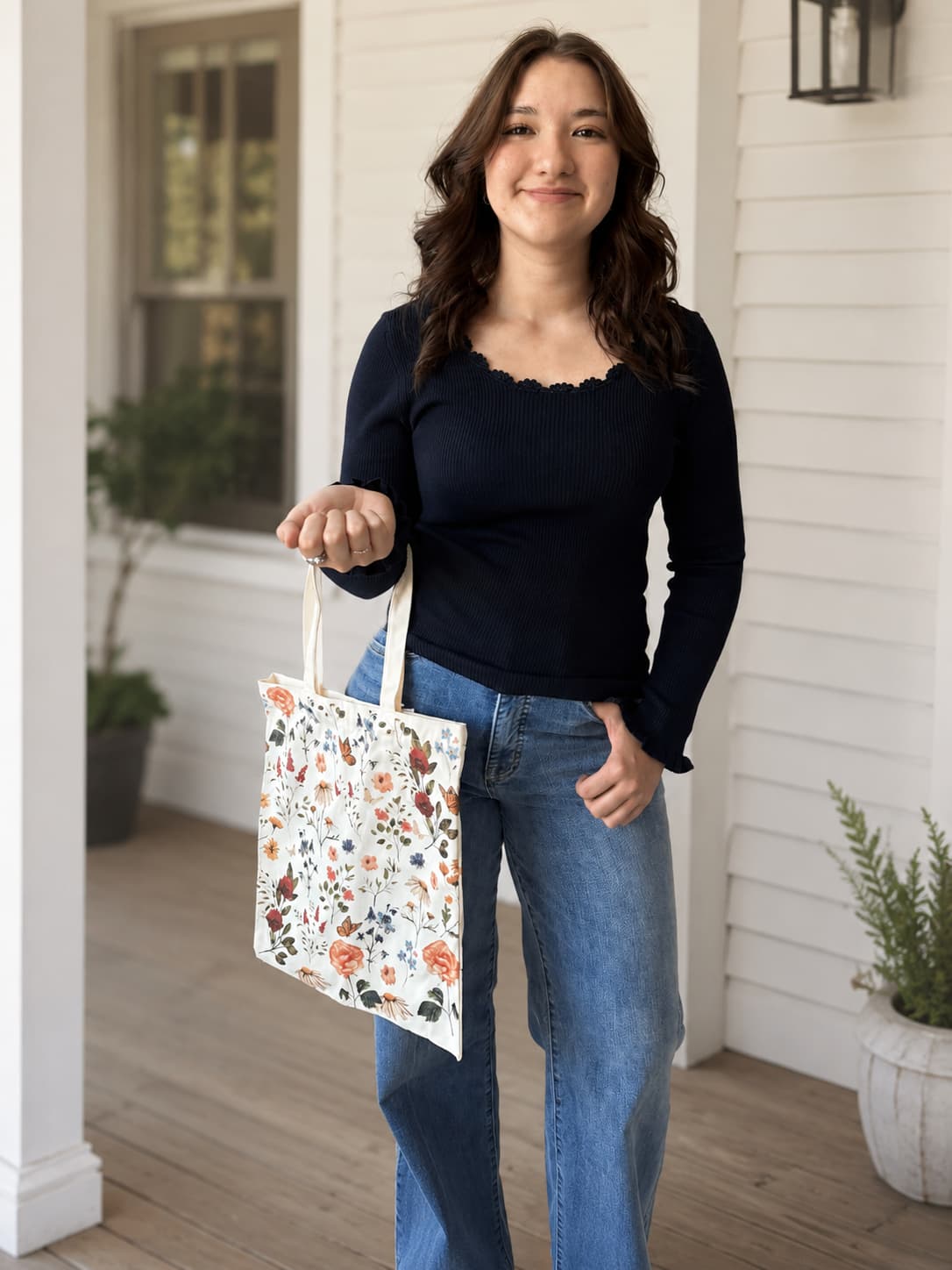 A woman stands on a white porch holding a white canvas tote bag decorated with a colorful floral and bird pattern. She is wearing a dark navy long-sleeve ribbed top with delicate neckline and ruffle wrist detail with blue wide leg jeans.