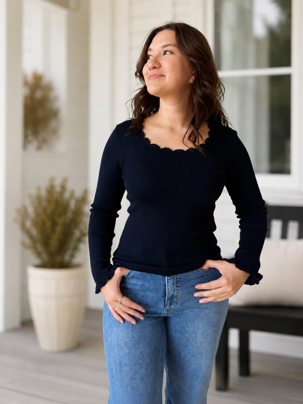 A woman wearing a dark navy long-sleeve ribbed knit top with a scalloped scoop neckline and ruffled cuffs, paired with blue jeans, standing on a porch.