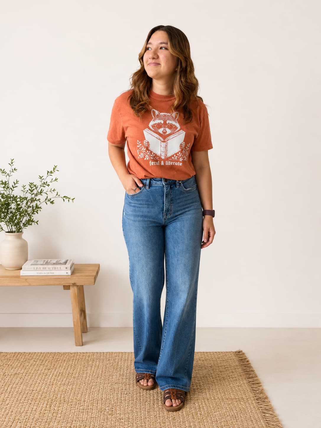 A woman stands indoors wearing a rust-orange graphic T-shirt with a white fox and book design, paired with blue jeans and brown sandals. She is posed on a woven rug beside a small wooden bench with a plant and books against a plain light wall.