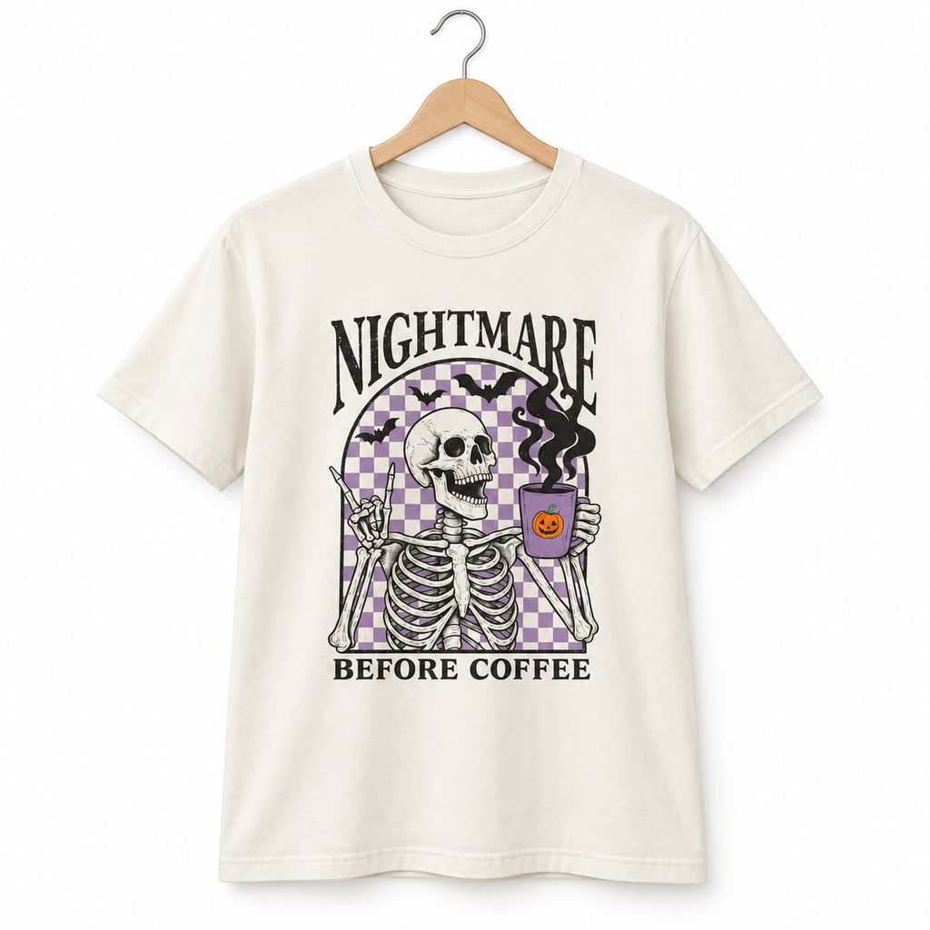 An off-white short-sleeve t-shirt hanging on a wooden hanger against a white background. The shirt features a graphic print of a skeleton holding a purple bucket with a jack-o'-lantern, with the text 'NIGHTMARE BEFORE COFFEE' in black.
