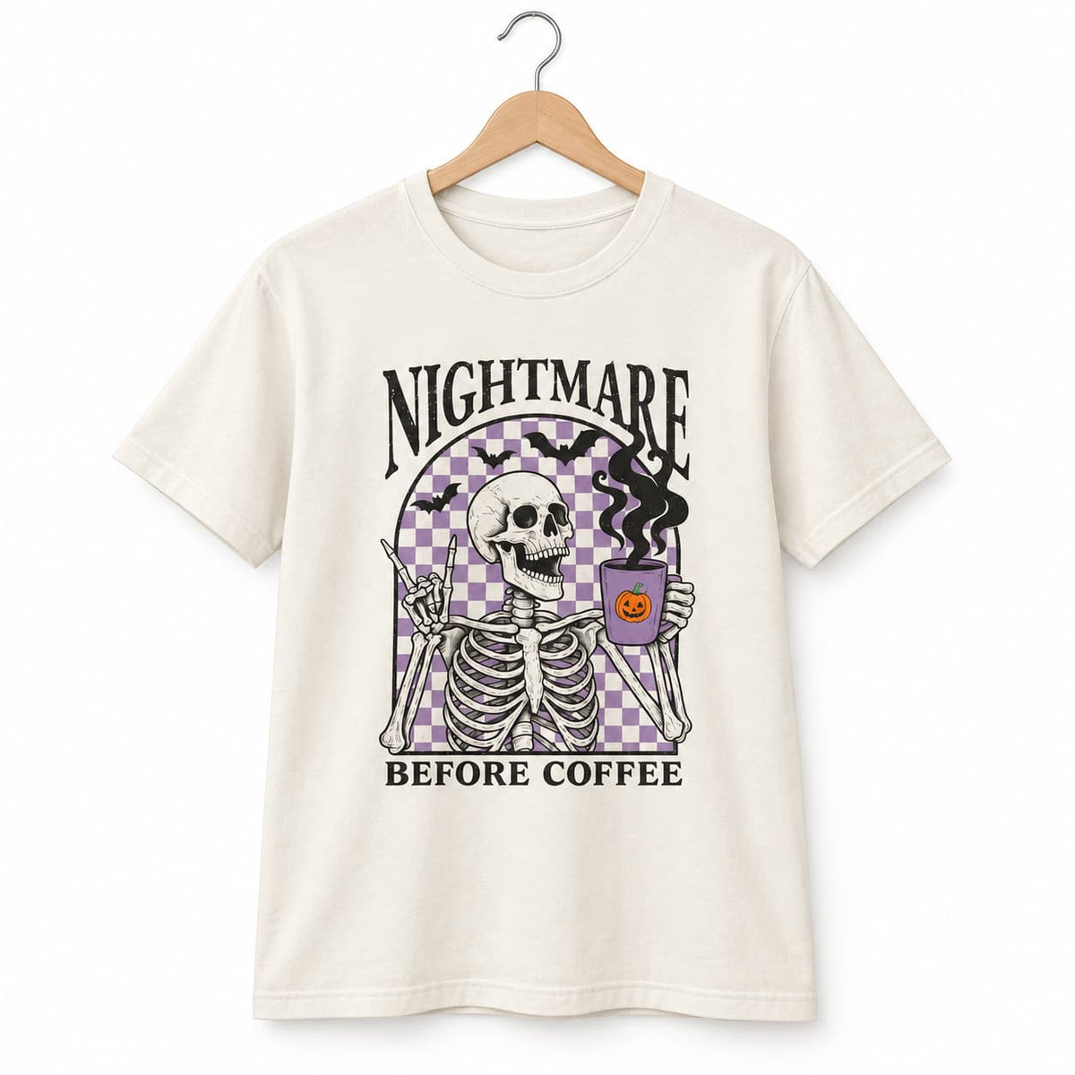An off-white short-sleeve t-shirt hanging on a wooden hanger against a white background. The shirt features a graphic print of a skeleton holding a purple bucket with a jack-o'-lantern, with the text 'NIGHTMARE BEFORE COFFEE' in black.