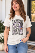 A woman wearing a cream-colored graphic t-shirt with a Halloween design featuring a skeleton and the text 'NIGHTMARE BEFORE COFFEE'.