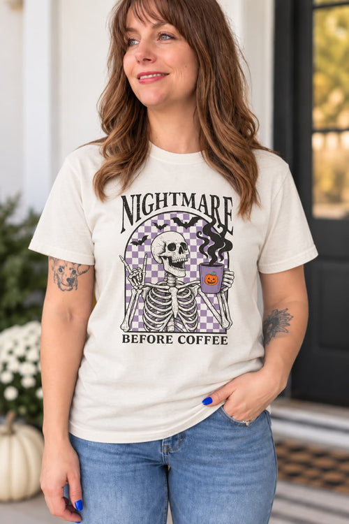 A woman wearing a cream-colored graphic t-shirt with a Halloween design featuring a skeleton and the text 'NIGHTMARE BEFORE COFFEE'.