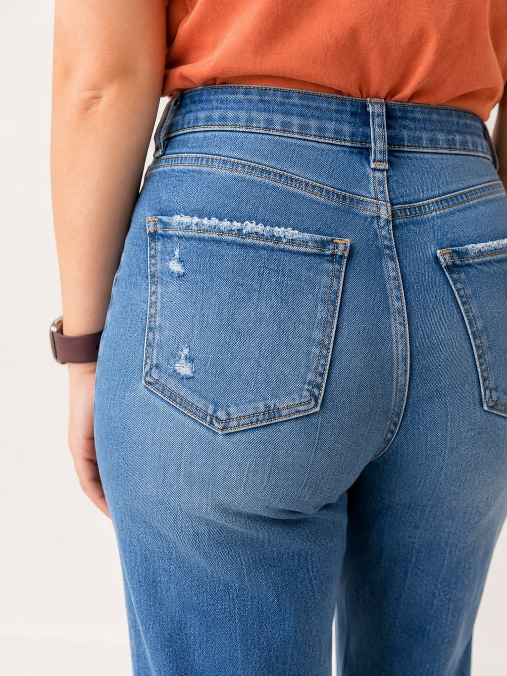 Close-up back view of a person wearing medium-wash blue denim jeans with distressed details on the back pocket, paired with an orange top against a plain light background.