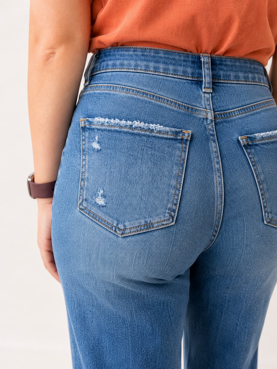 Close-up back view of a person wearing medium-wash blue denim jeans with distressed details on the back pocket, paired with an orange top against a plain light background.