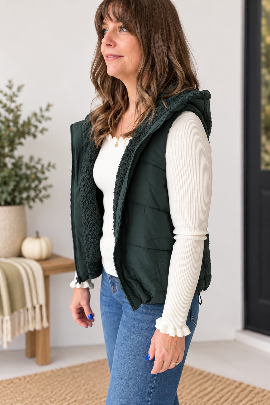 A woman wearing a dark forest green hooded puffer vest with a textured sherpa lining over a white ribbed long-sleeve top and blue jeans, standing in a brightly lit indoor setting.