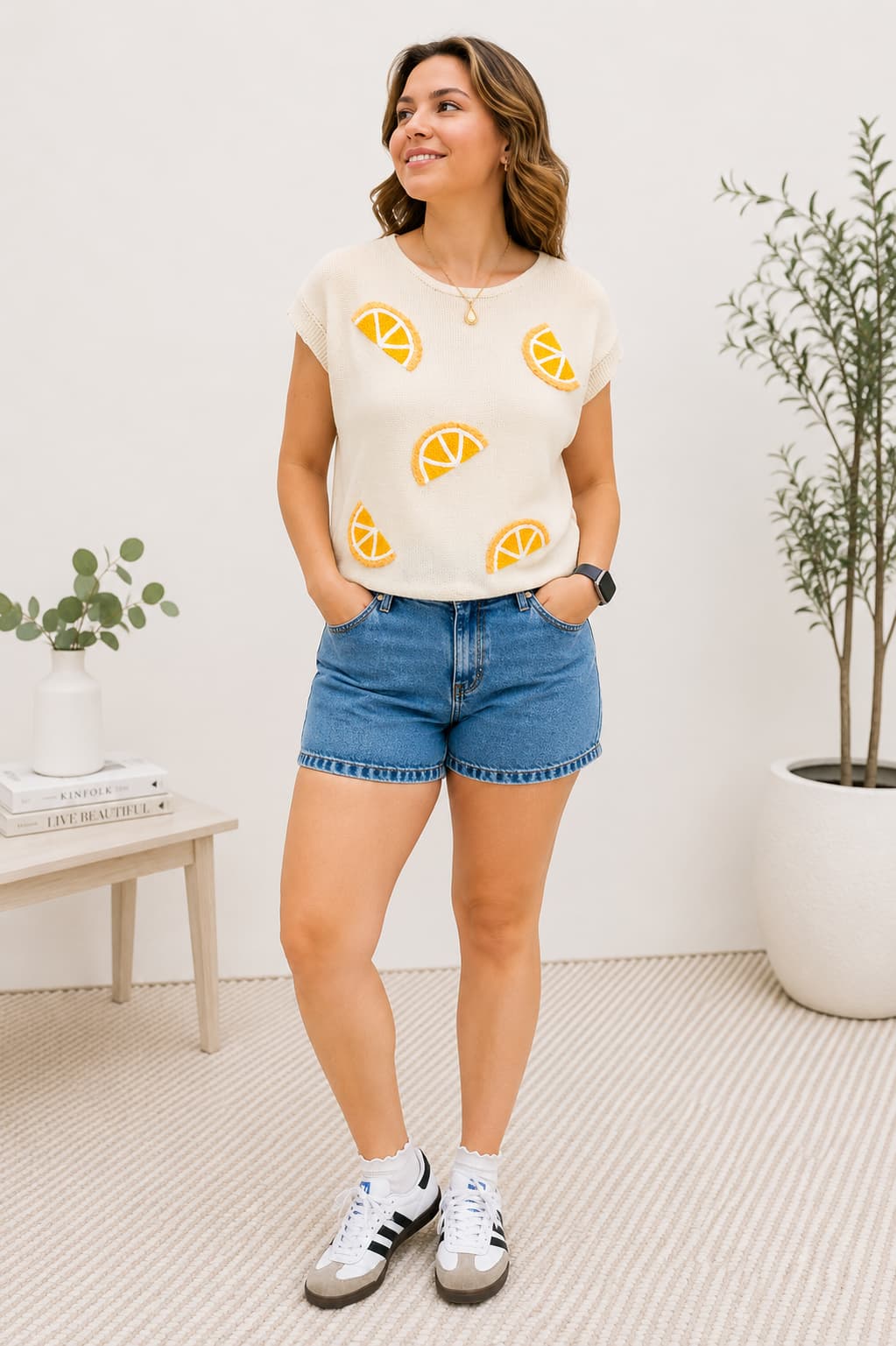 Woman wearing a knit top with orange designs and blue shorts, standing in a room with plants.