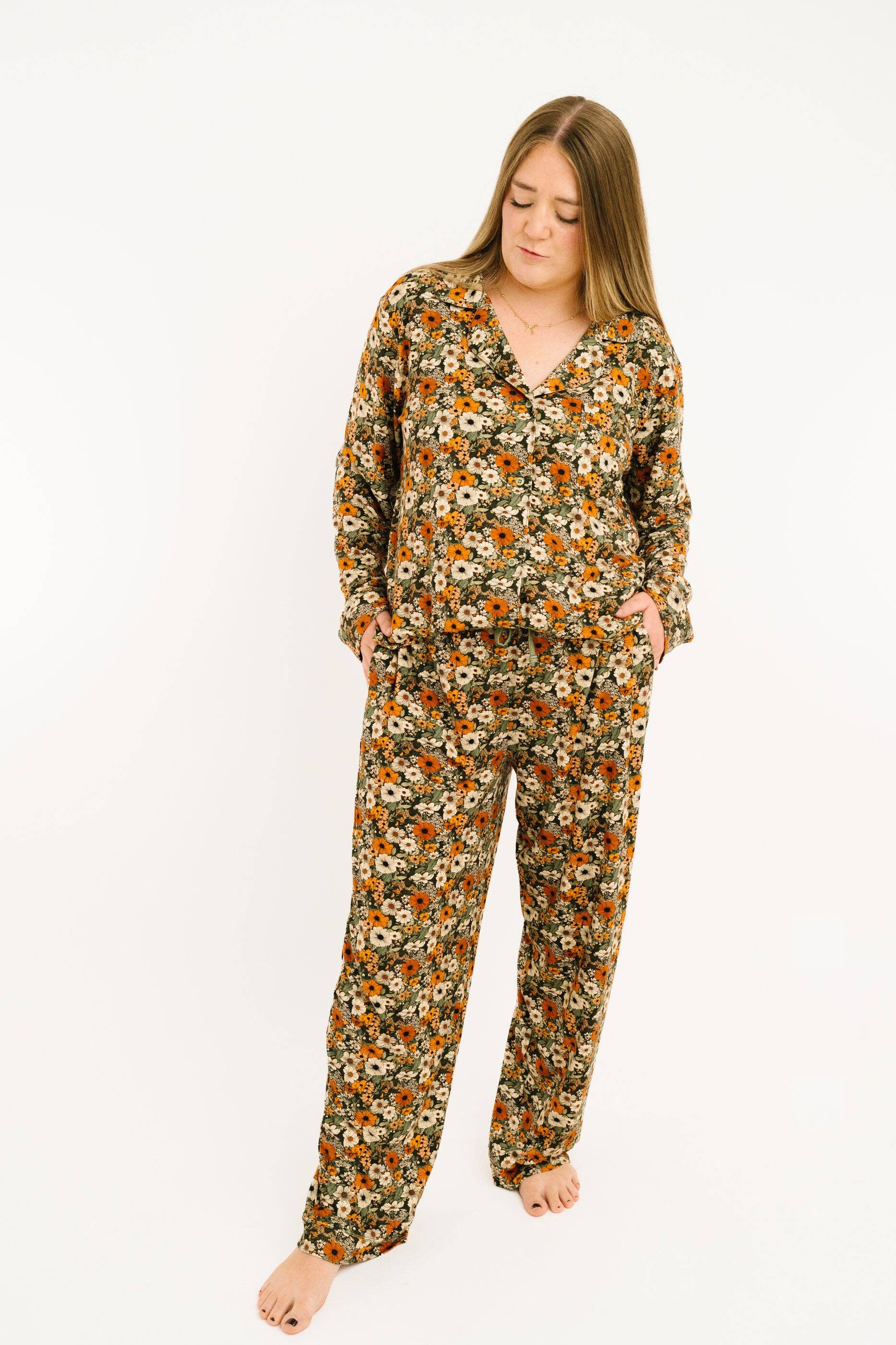 A woman modeling a two-piece long-sleeve pajama set with an all-over floral print in orange, yellow, white, and green on a dark background, photographed against a plain white studio background.