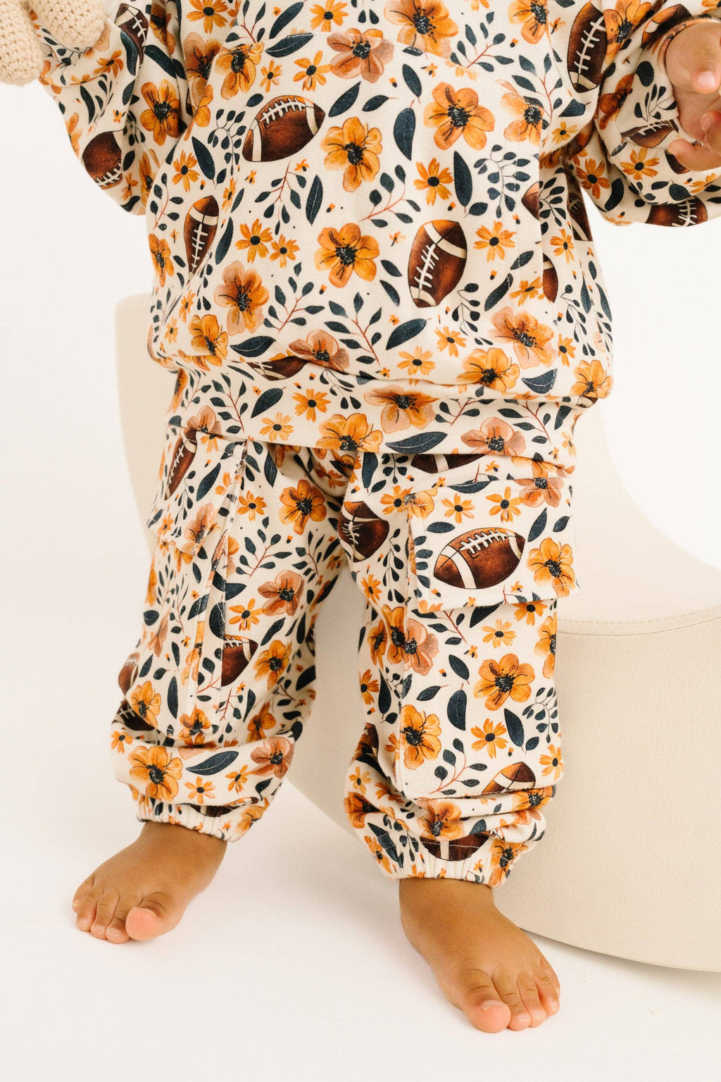 A toddler wearing a matching two-piece cotton set with a football-themed floral print, featuring orange and yellow flowers, green leaves, and brown football motifs on an off-white background.