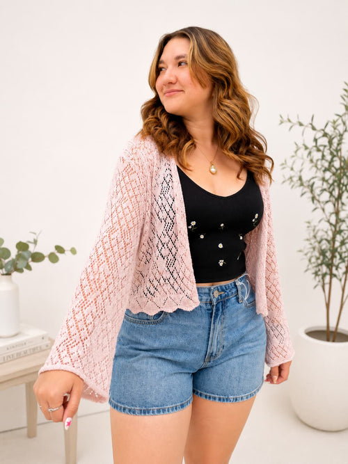 Woman wearing a pink knit bell sleeve cardigan over a black top and denim shorts  indoors.