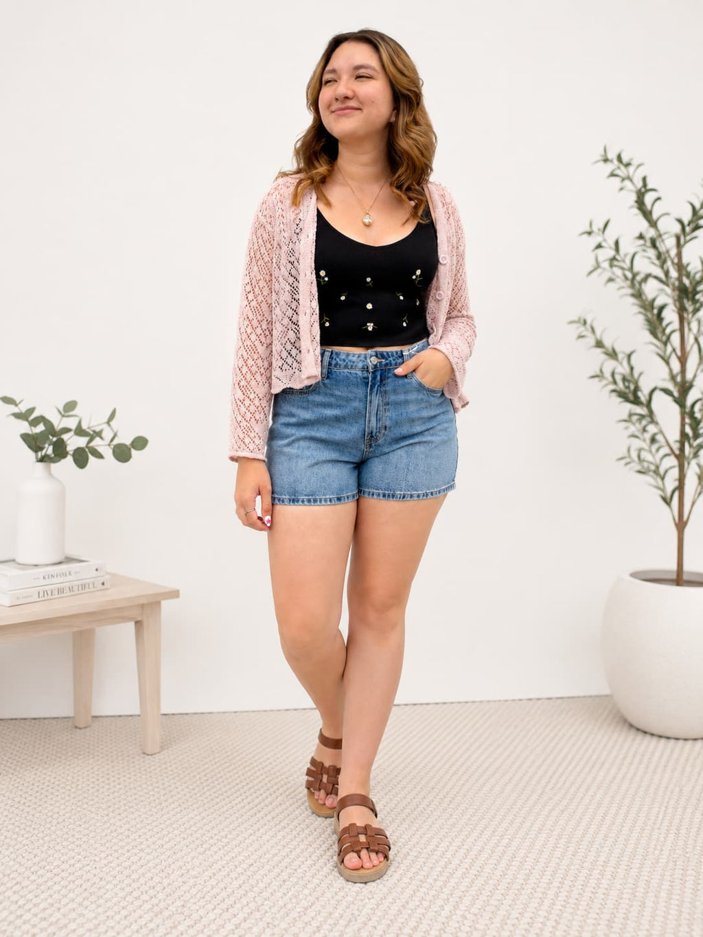 Woman wearing a black top, pink cardigan, and denim shorts standing in a room with white walls and a small table.