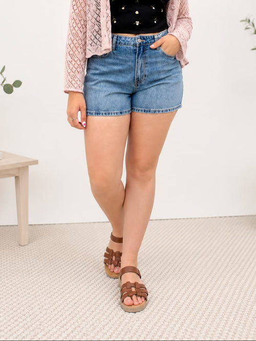 Woman wearing a black top, pink cardigan, and denim shorts standing in a room with white walls and a small table.