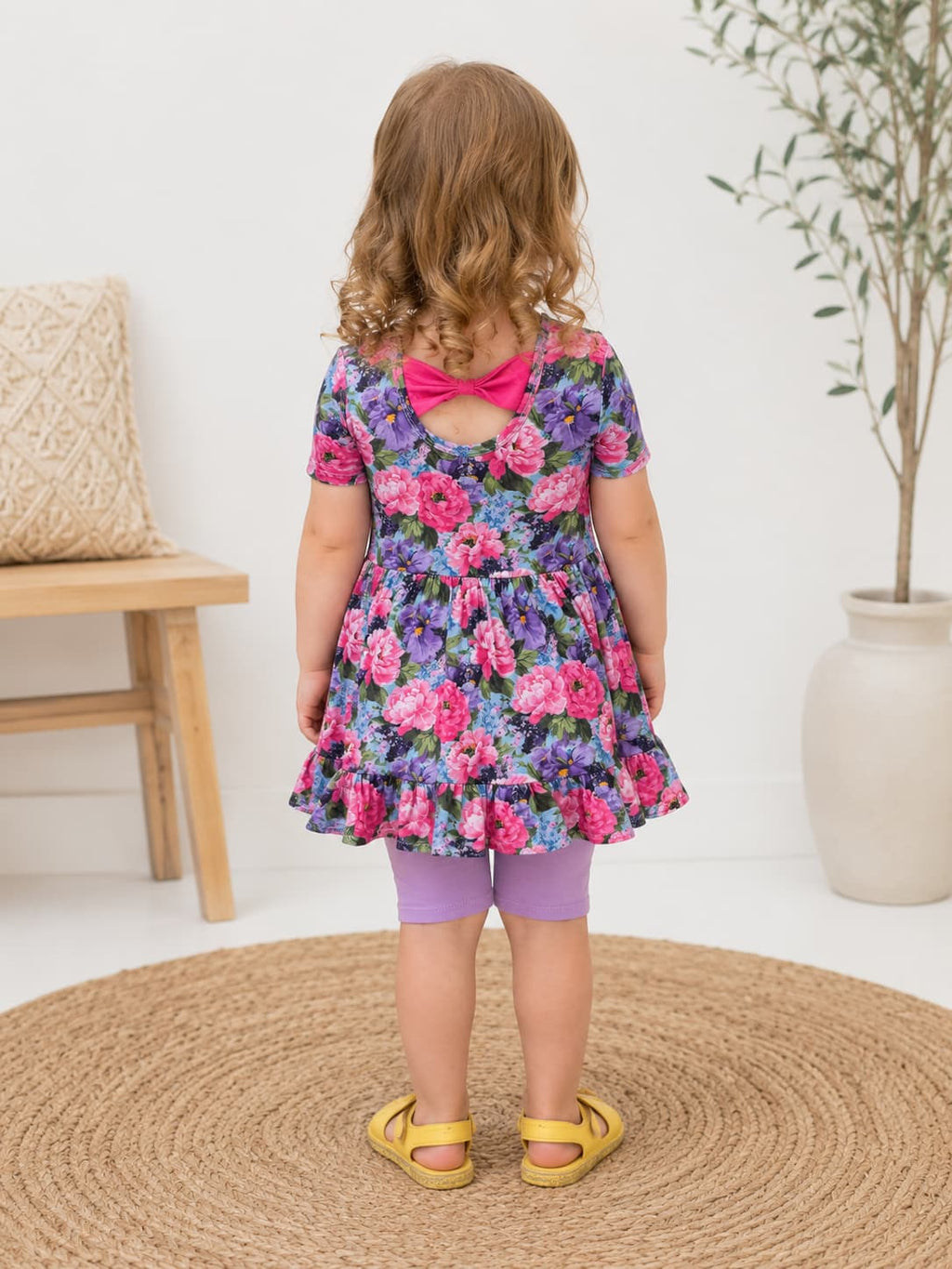 A toddler girl is shown from behind wearing a short-sleeved floral dress with a bright pink bow at the back neckline, purple leggings, and yellow sandals, standing on a round woven rug in a bright room.