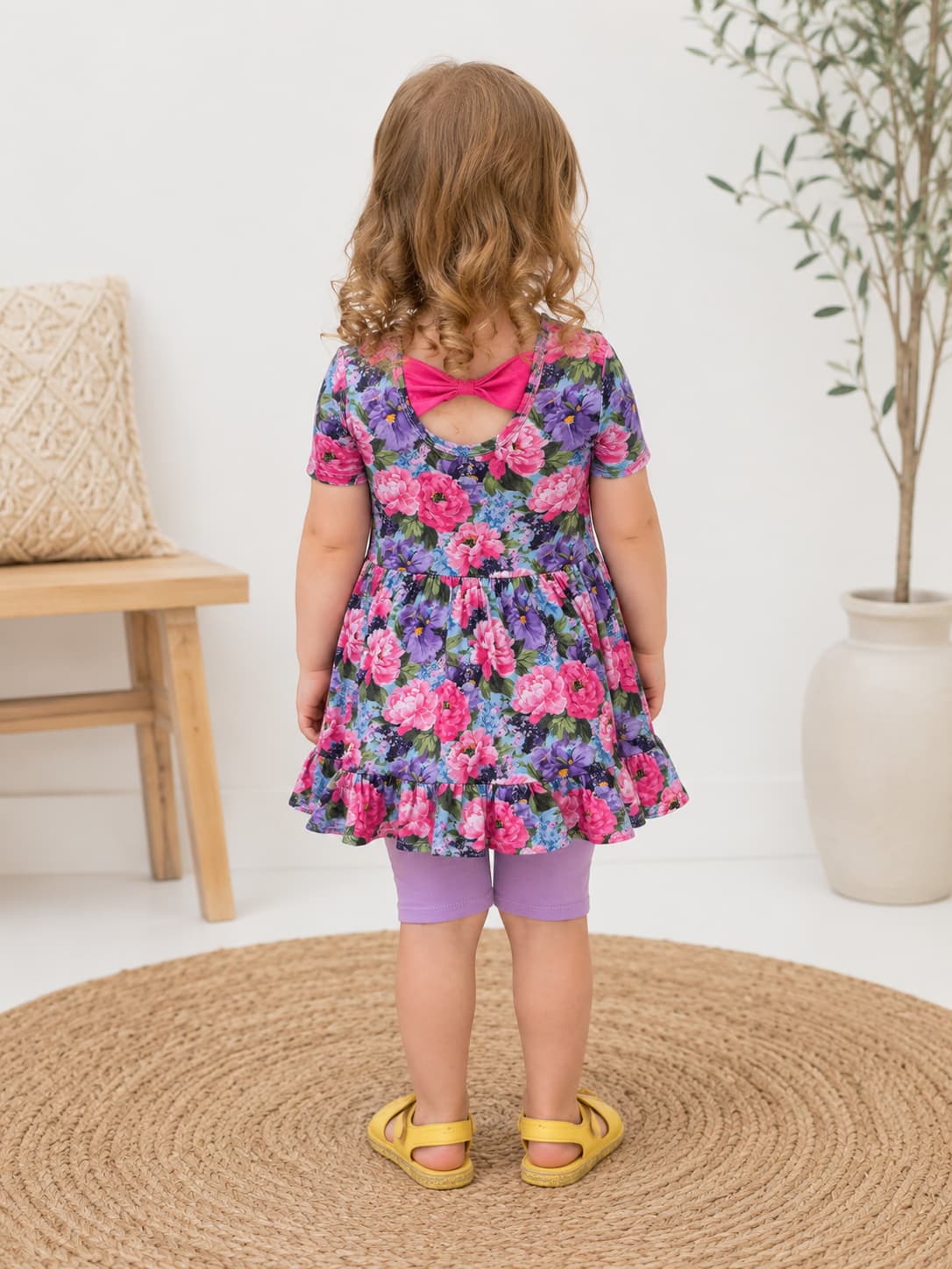 A toddler girl is shown from behind wearing a short-sleeved floral dress with a bright pink bow at the back neckline, purple leggings, and yellow sandals, standing on a round woven rug in a bright room.