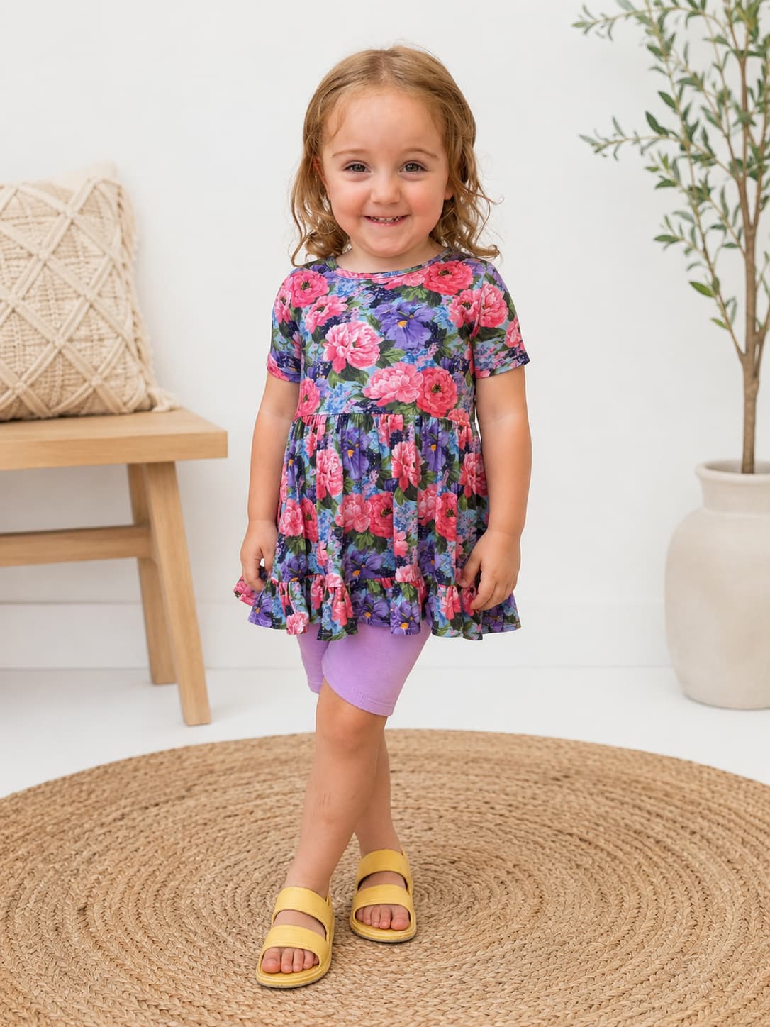 A young girl stands on a round woven rug wearing a short-sleeve floral dress with purple ringer shorts and yellow sandals in a bright indoor room.