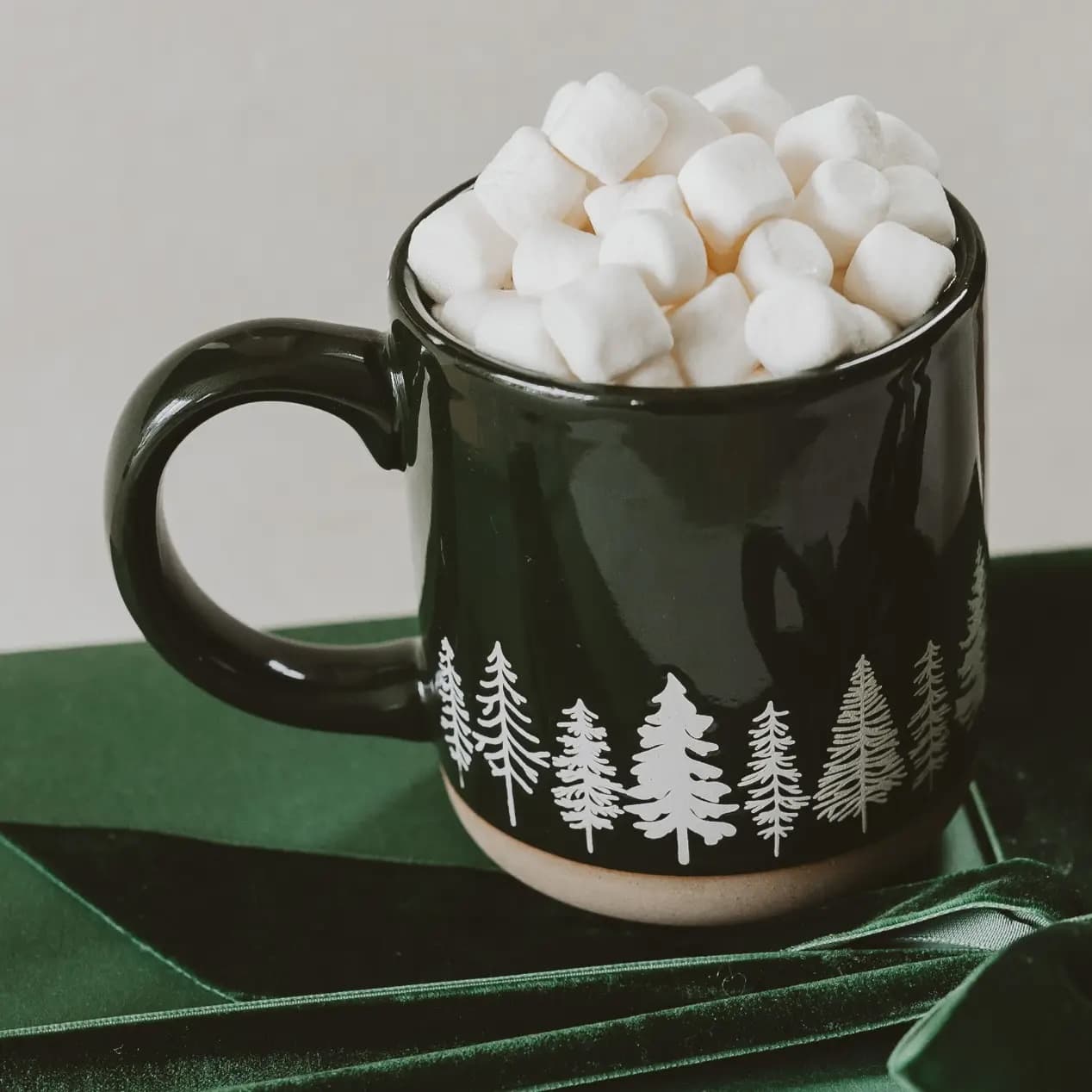 A glossy dark green ceramic mug filled with mini marshmallows, decorated with a white pine tree pattern near the base, set on a dark green ribbon against a neutral background.