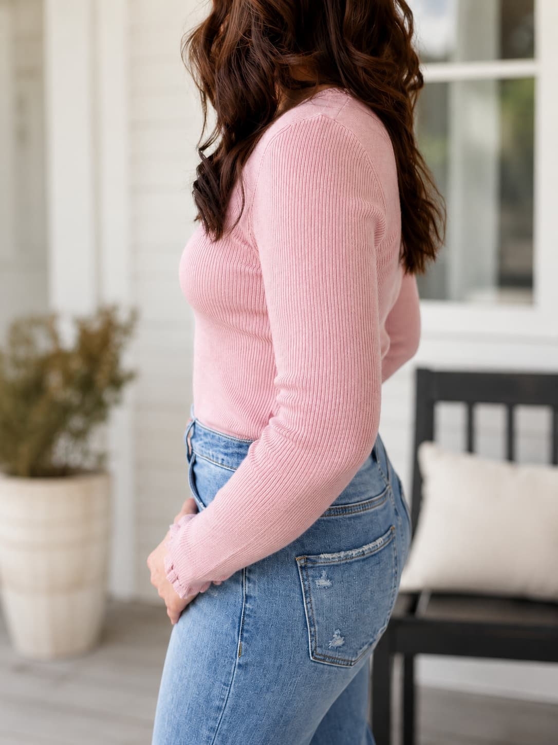 A woman shown from the side wearing a light pink ribbed long-sleeve top and light blue jeans, standing on a porch with a chair and potted plant in the background.