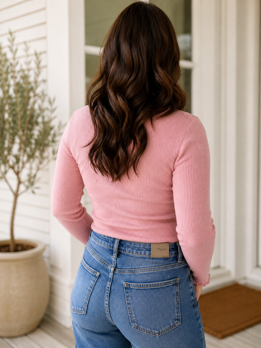 A woman viewed from behind wearing a pink ribbed long-sleeve top and blue jeans, standing in front of a white door with a potted plant to the left.