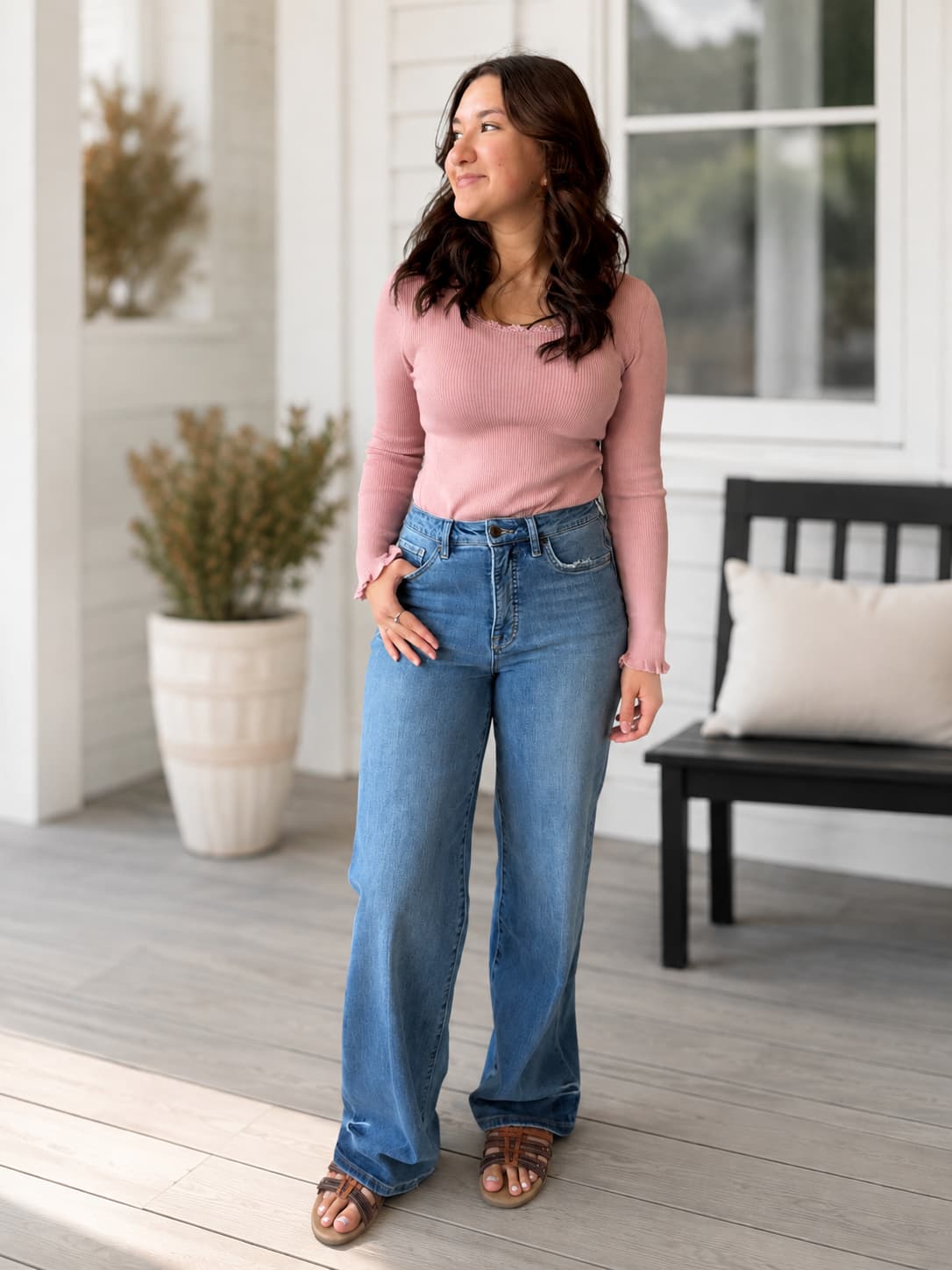 A woman stands on a porch wearing a dusty pink long-sleeve knit top with delicate scallop neckline, ruffle wrist detail and high-waisted, wide-leg blue jeans. She is wearing brown leather slide sandals.