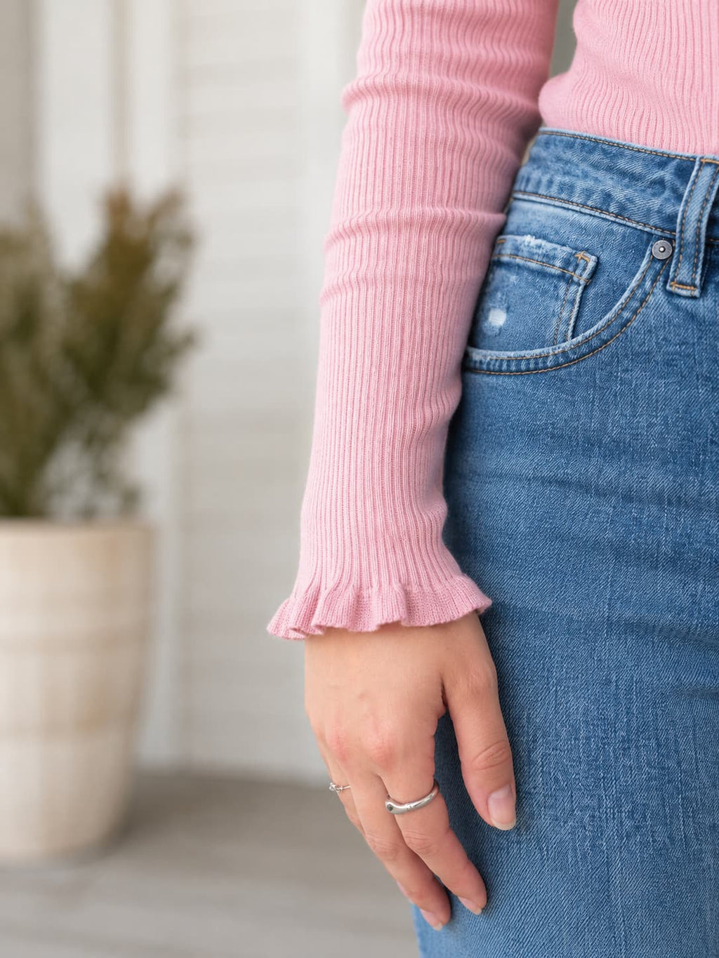 Close-up of a person wearing a light pink ribbed long-sleeve top with a ruffled hem and blue denim jeans, standing indoors with a blurred potted plant in the background.