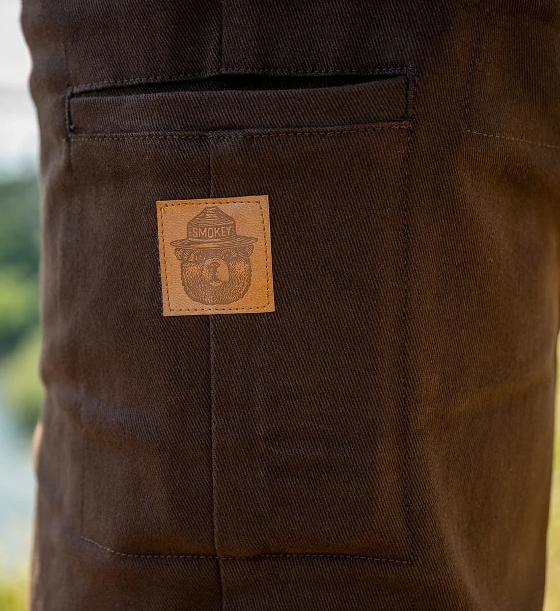 Close-up of dark brown canvas pants showing a front pocket with a tan leather patch with Smokey the Bears face that reads 'SMOKEY'.