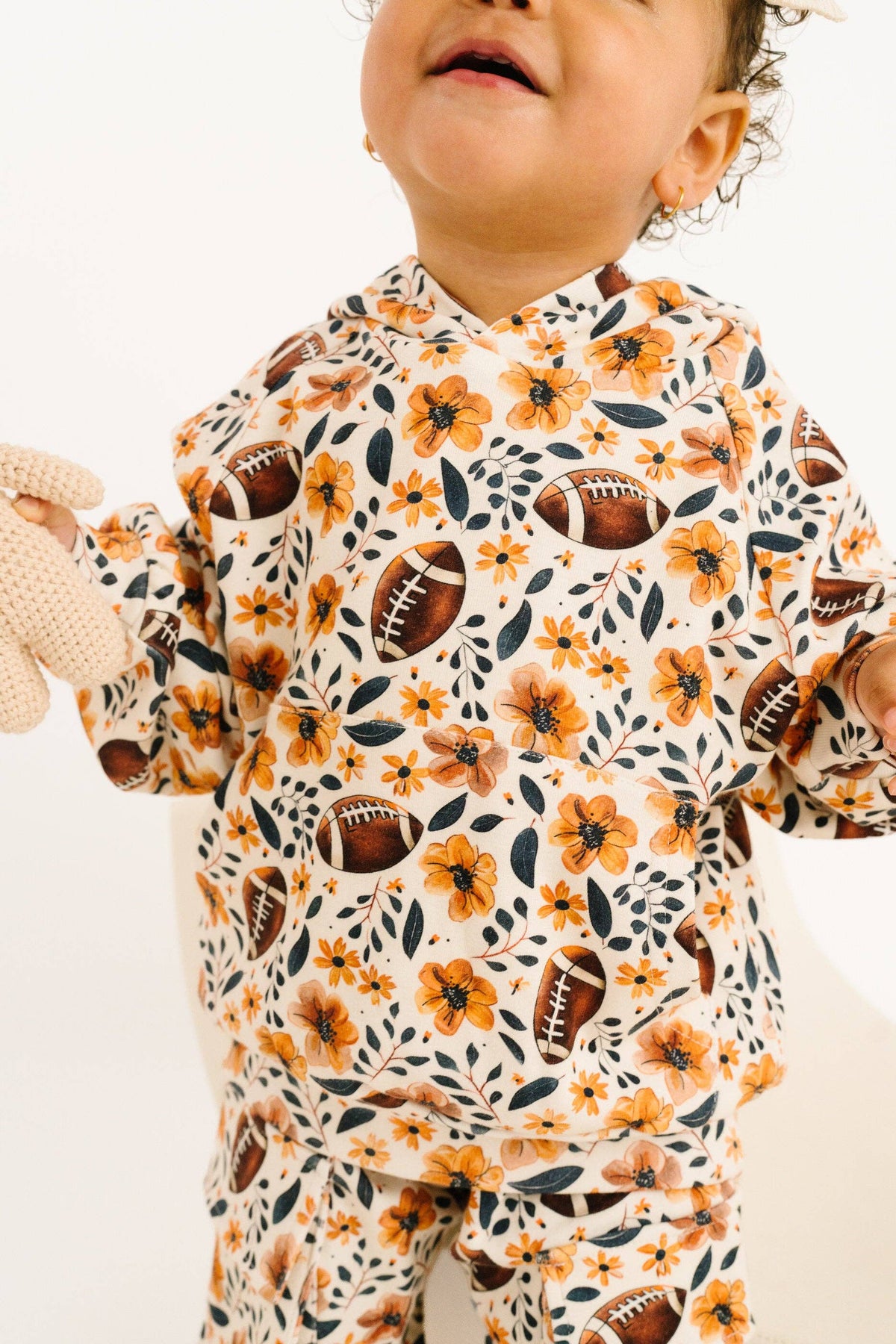 A close-up of a baby wearing a long-sleeve hooded two-piece outfit with an all-over autumn floral and football print on a cream background.