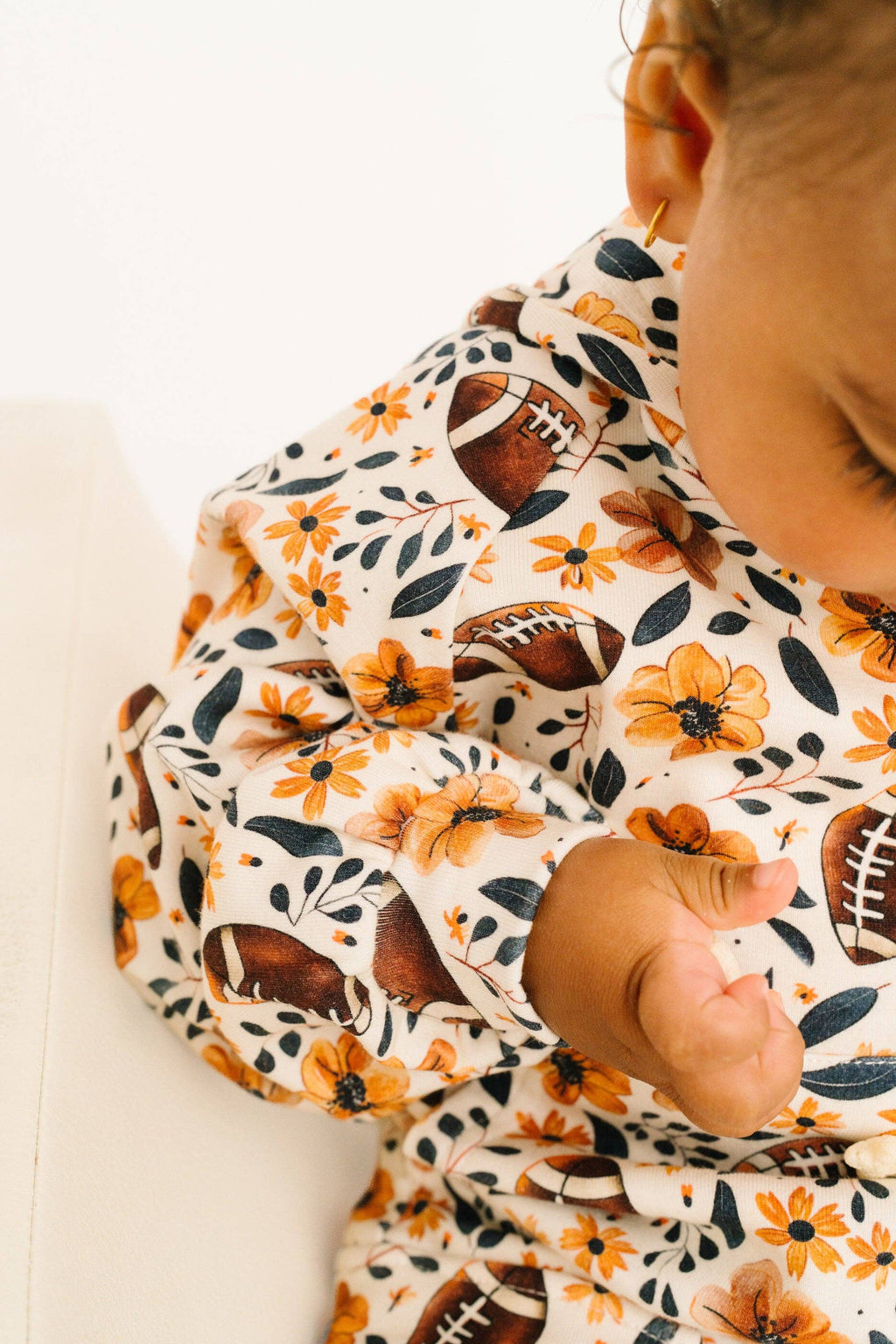 A close-up lifestyle shot of a baby wearing a cream-colored long-sleeve top with a pattern of orange flowers, dark green leaves, and brown footballs.