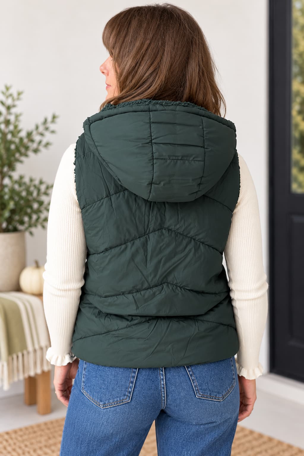 A woman shown from the back wearing a dark green hooded puffer vest over a cream ribbed long-sleeve top and blue jeans in a bright indoor setting.