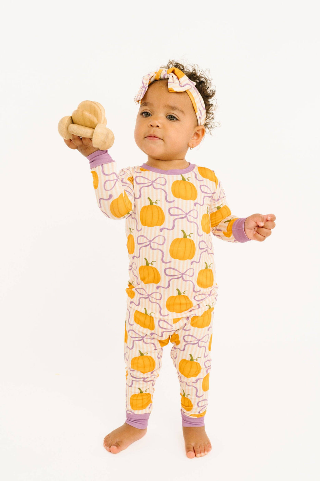 A baby standing barefoot against a white background, wearing a long-sleeve pajama romper with an orange pumpkin and purple ribbon pattern, a matching headband, and holding a small stuffed animal.