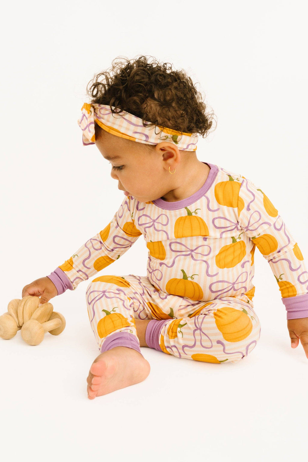 A baby wearing a long-sleeve pajama set and matching headband with a pumpkin and bow pattern, sitting on a white background and playing with a wooden toy.