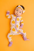 A baby lying on an orange background wearing a one-piece pajama with a pumpkin print and purple trim, along with a matching headband.