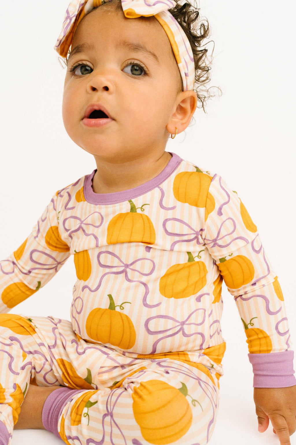 A baby wearing a long-sleeve pajama top and matching pants with a pumpkin and purple bow pattern, along with a matching headband, photographed against a plain white background.