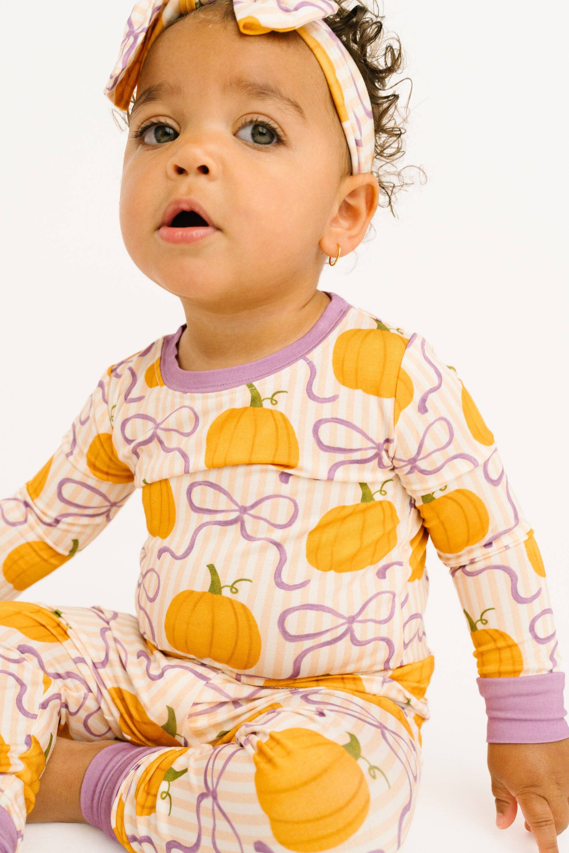 A baby wearing a long-sleeve pajama top and matching pants with a pumpkin and purple bow pattern, along with a matching headband, photographed against a plain white background.