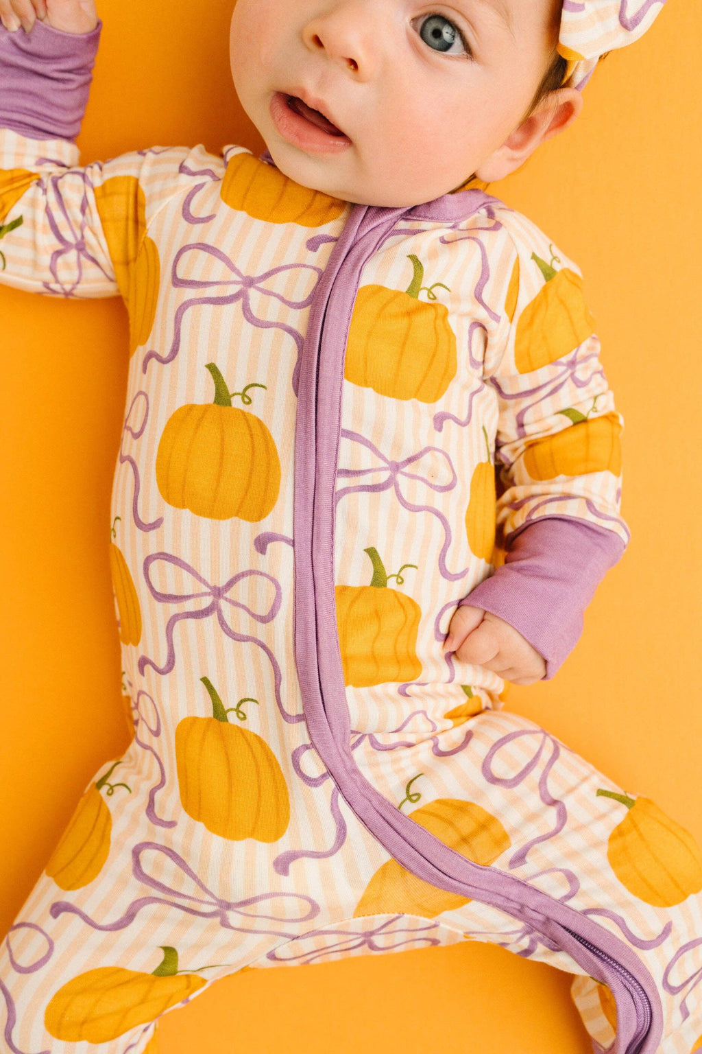 A baby wearing a footless zip-up sleeper with a pumpkin and bow pattern, lying on a solid orange background.