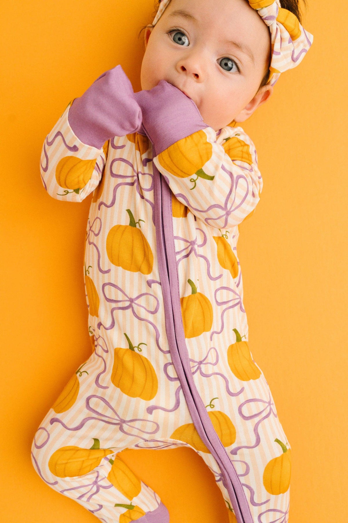 A baby wearing a footed one-piece sleeper with a pumpkin and ribbon pattern, lying on a solid orange background. The sleeper has purple trim and a matching headband.
