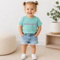 A toddler girl stands indoors wearing a mint green graphic T-shirt with pink text reading 'Put it on my dad's tab', light blue denim shorts, and white sneakers. The room has a white wall, a woven pouf, a small wooden side table, and a potted plant.