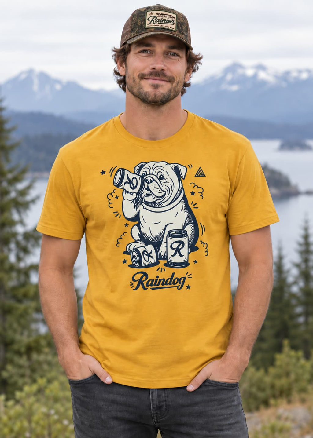 A man wearing a mustard yellow graphic t-shirt with a cartoon bulldog and beer cans design, paired with a camouflage cap, standing outdoors in front of a mountain and lake background.
