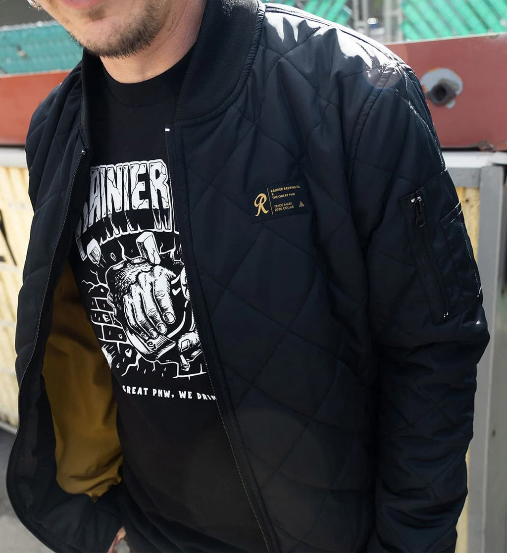 A man wearing a black quilted bomber jacket over a black graphic t-shirt and a brown shirt, photographed outdoors.