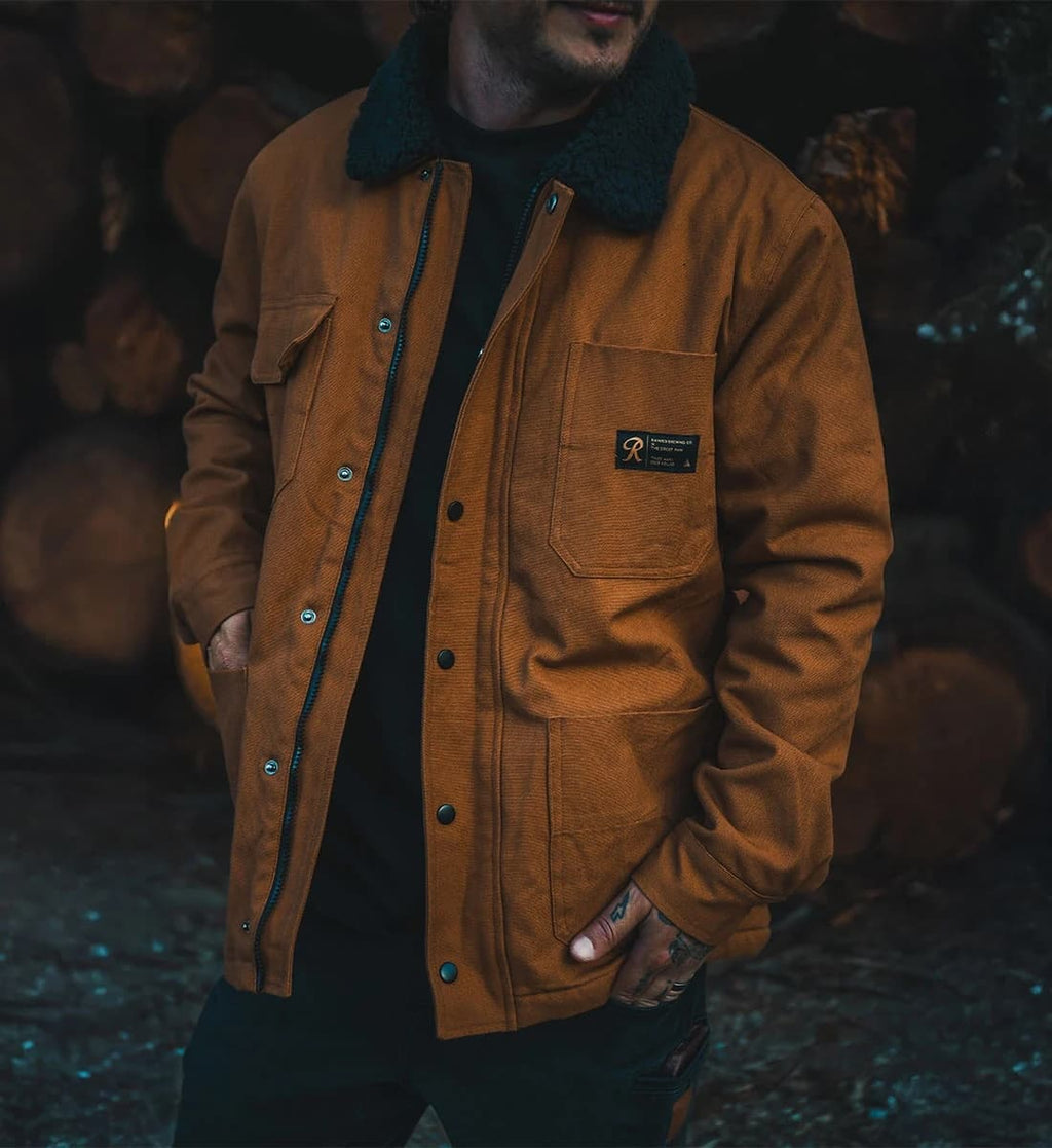 A man wearing a brown waxed canvas utility jacket with a dark fleece collar, standing in front of a dark background of stacked logs.